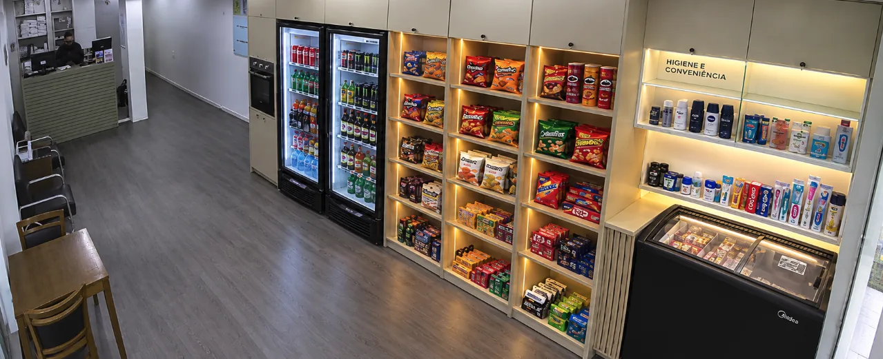 Interior da conveniência, com geladeiras de bebidas, prateleiras iluminadas de snacks e doces, área de higiene e freezer de congelados